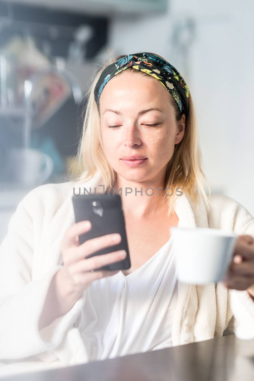 Beautiful caucasian woman at home, feeling comfortable wearing white bathrobe, taking some time to herself, drinking morning coffee and reading news on mobile phone device in the morning by kasto