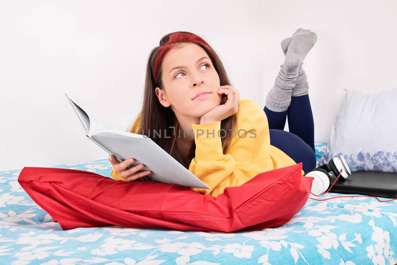 Beautiful young girl in her room holding a book, looking to the side, thinking, wondering, fantasizing while lying in bed next to laptop and headphones.