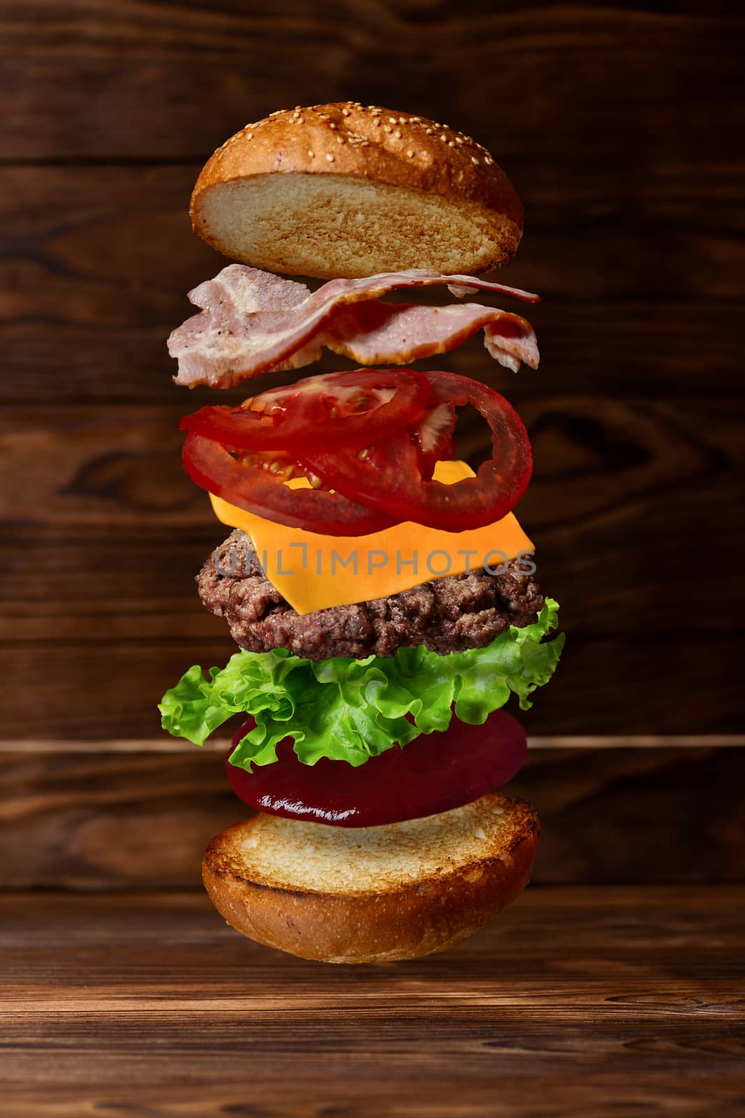 Big tasty burger with flying ingredients on wooden background. Stock photo of food levitation. Includes bun, sauce, salad leaf, cutlet, cheese, tomato slices and bacon