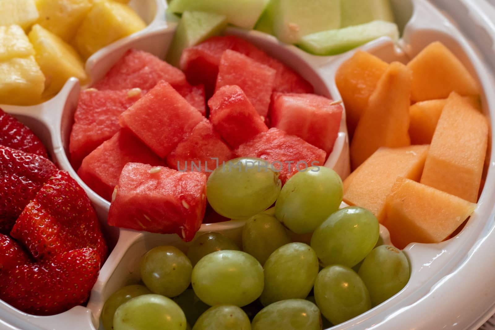 A platter of fruit, consisting of watermelon, cantaloupe, green grapes, strawberries, pineapple and honeydew melon is served in a white plastic tray as a snack.