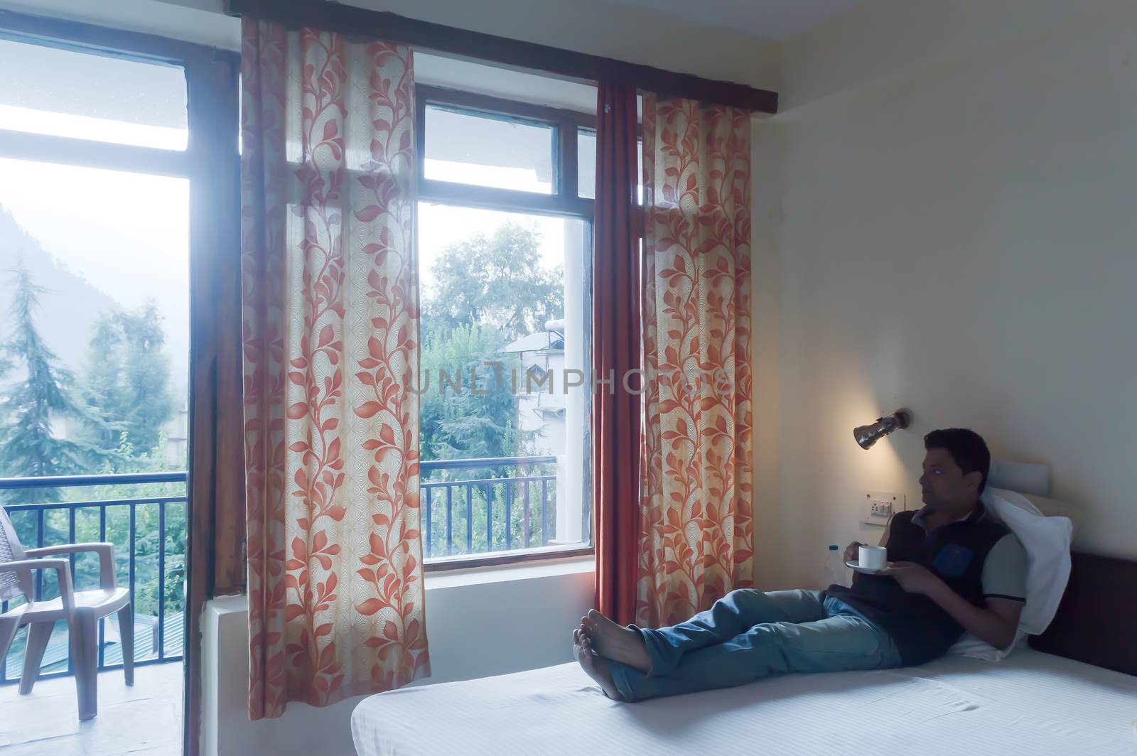 Mid adult man solo business Traveler of Indian ethnicity in casual clothing, laying on bed of a holiday villa and holding a hot cup of tea in morning looking outside Mountain city background. Travel vacation active life and healthy lifestyles concept. by sudiptabhowmick