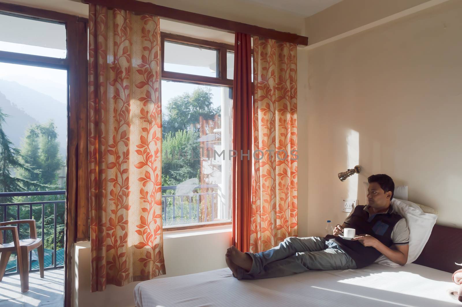 Mid adult man solo business Traveler of Indian ethnicity in casual clothing, laying on bed of a holiday villa and holding a hot cup of tea in morning looking outside Mountain city background. Travel vacation active life and healthy lifestyles concept. by sudiptabhowmick