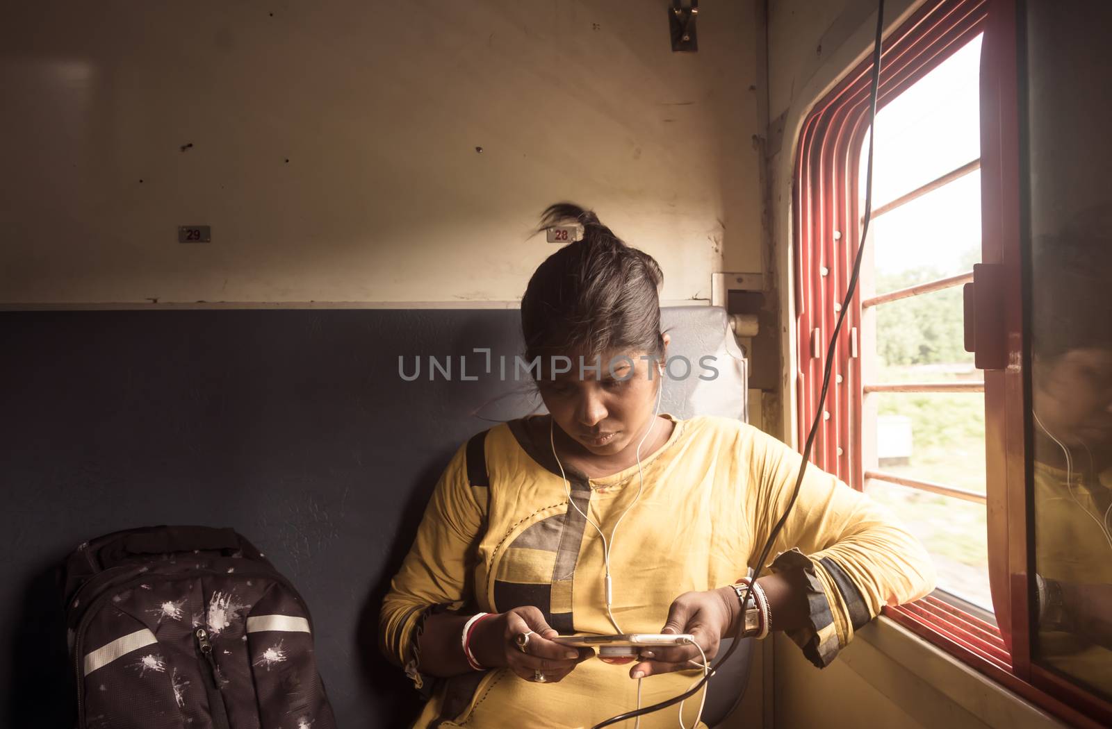 Woman using mobile phone while traveling solo in passenger train. Traveler enjoying Modern technology on the move in everyday life and travel. Close up portrait - Young Adult Lady - Indian ethnicity.