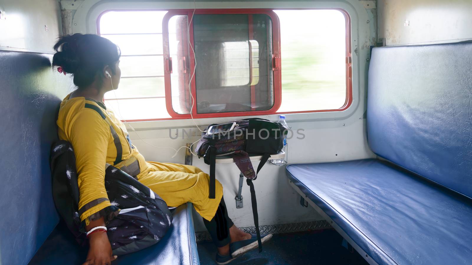 Woman listening music on mobile phone and looking outside from window while travelling solo in train. Traveler enjoying Modern technology on the move in everyday life and travel. Side view Close up portrait - Young Adult Lady - Indian ethnicity. by sudiptabhowmick