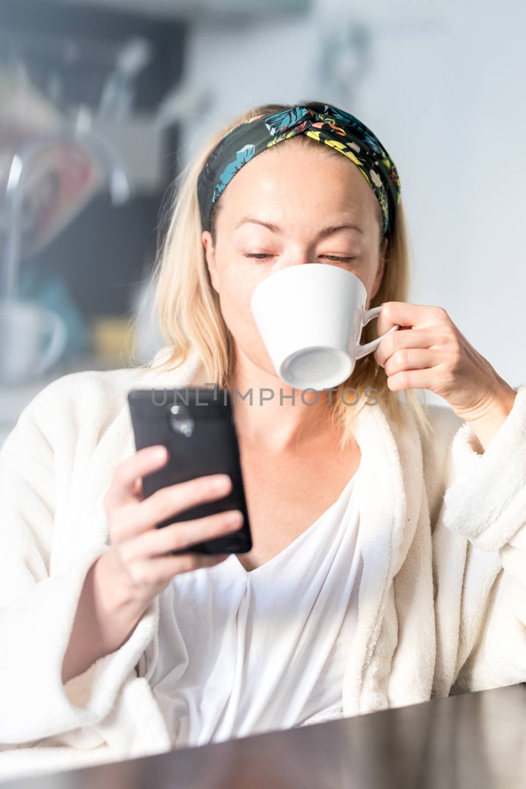 Beautiful caucasian woman at home, feeling comfortable wearing white bathrobe, taking some time to herself, drinking morning coffee and reading news on mobile phone device in the morning.