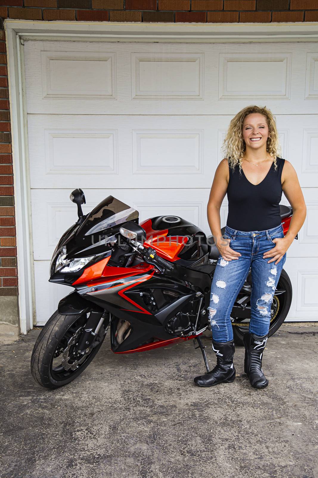 Twenty something blond woman wearing stylish ripped jeans and a black tank top, standing in from of a sport motocycle, with a happy expression