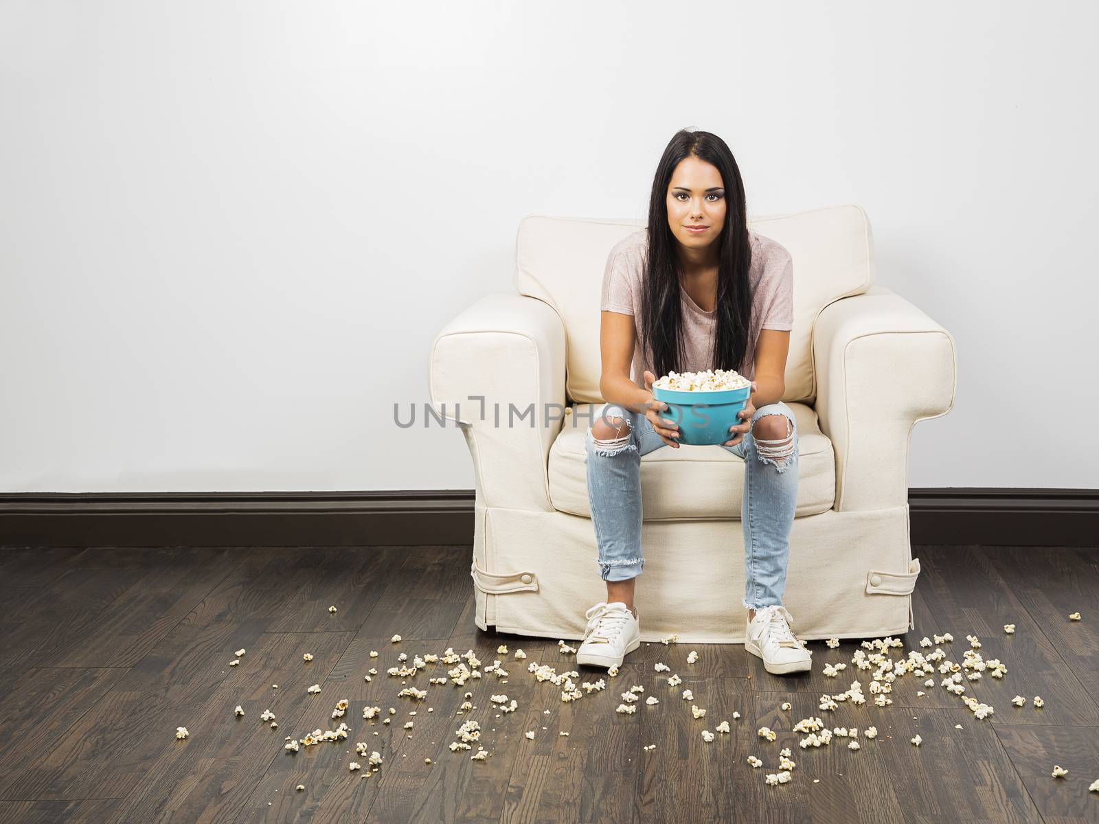 Young woman sitting on white couch holding a blue bowl with popcorn on the floor
