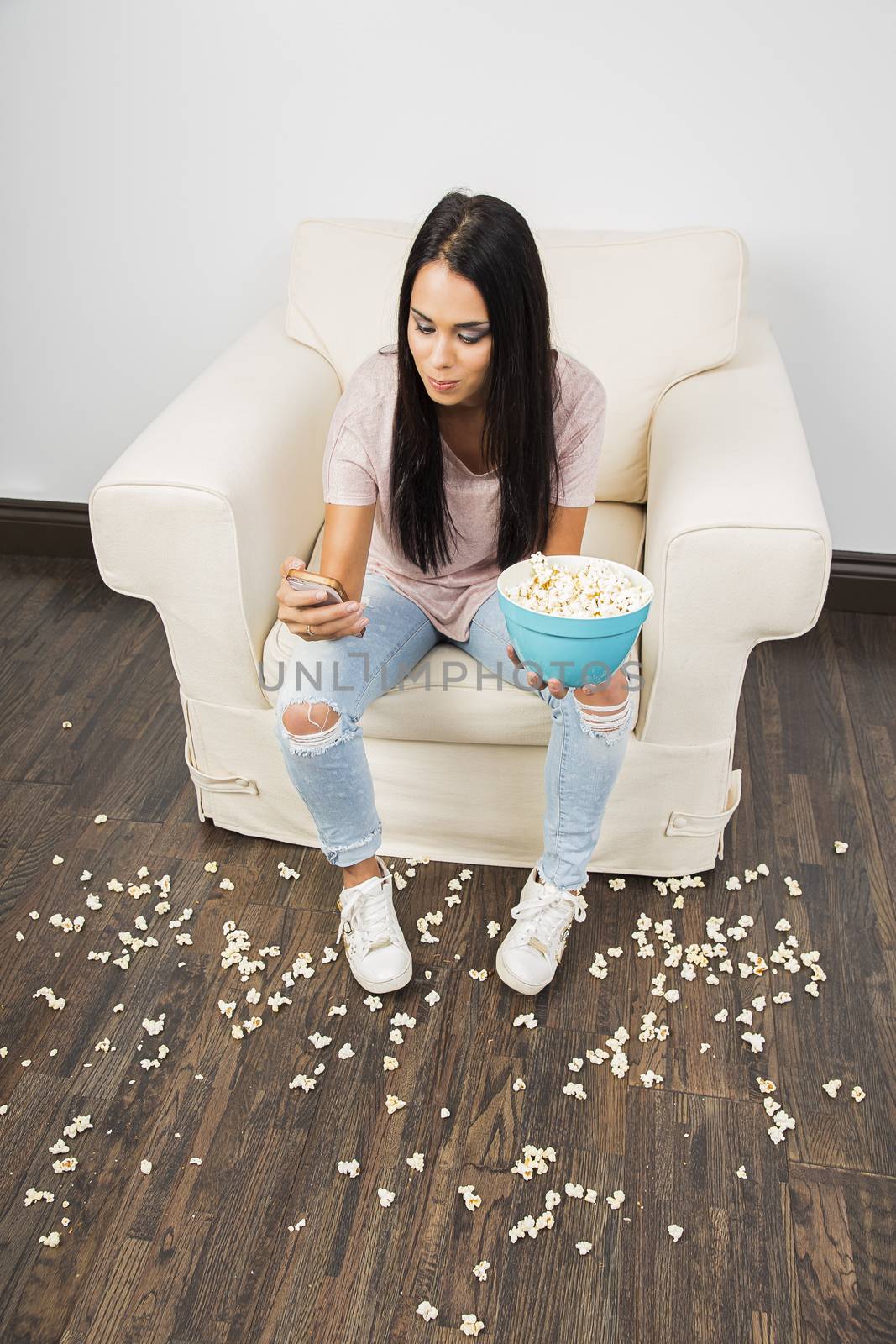 Young woman sitting on white couch holding a blue bowl, with popcorn on the floor, looking at her cell phone
