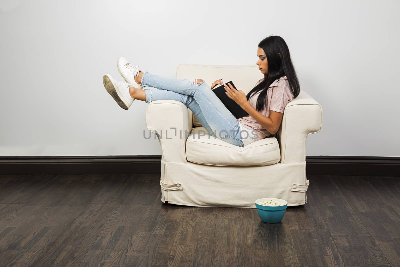 young woman, sitting on a white couch, reading a book with a bowl of popcorn