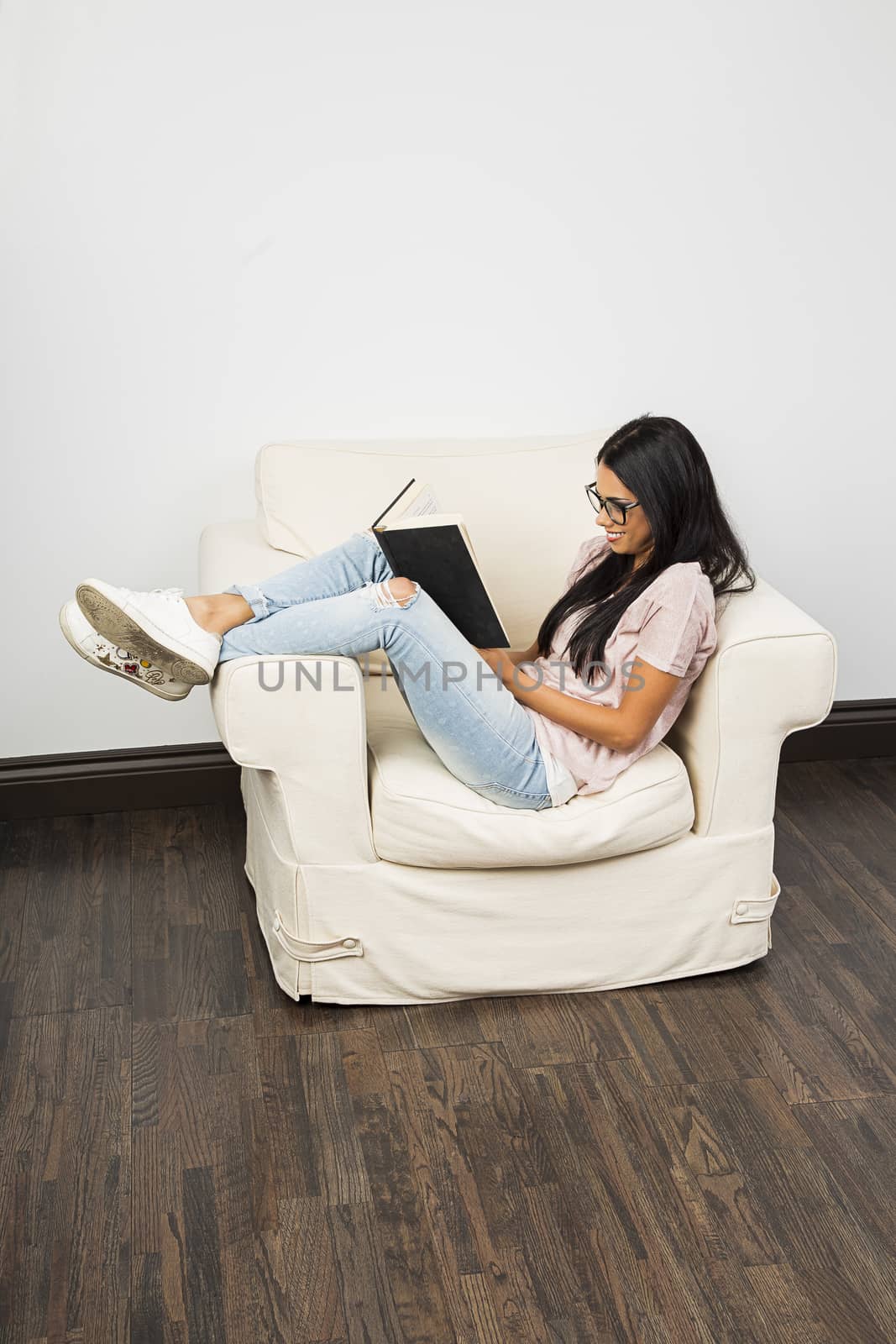 young woman wearing glasses, with her leg up on the arm of a couch, reading a hard cover book