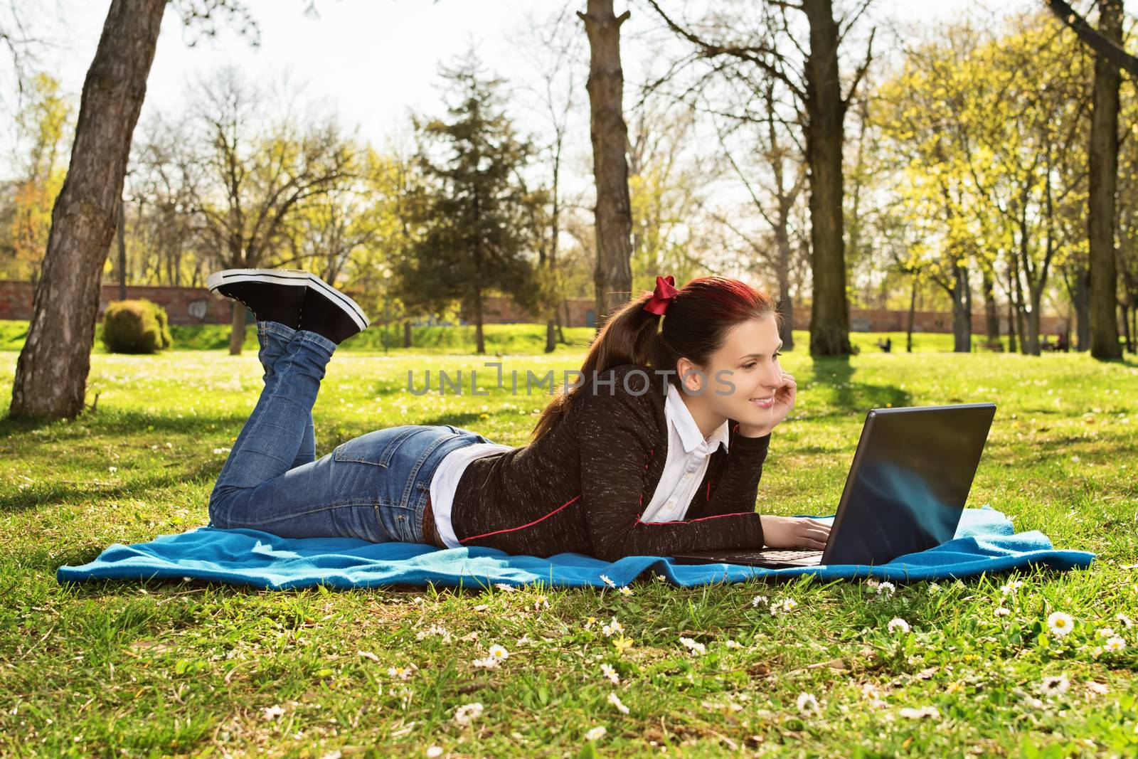 Beautiful casual student girl with laptop outdoors. Smiling woman lying on a blanket on the grass with computer, surfing on the Internet or preparing for exams. Technology, education and remote working concept.

