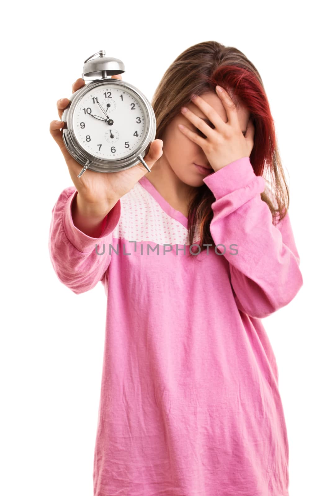 Being late concept. Close up of a young woman in pink pajamas holding an alarm clock and covering her face with her hand, looking tired and annoyed, isolated on white background.