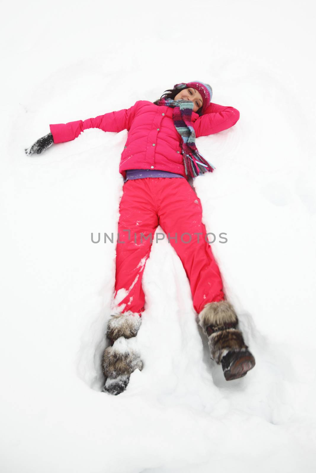 Beautiful joyful young woman in pink clothes laying down on a snow on vacation during a sunny winter day