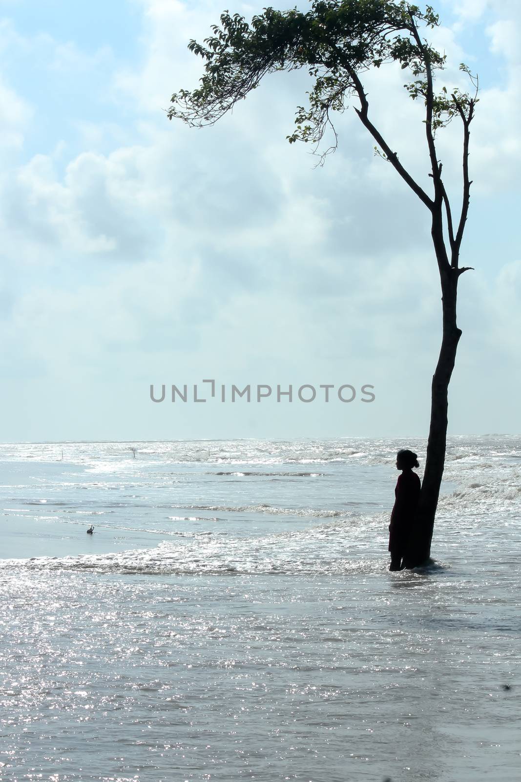 A solitary lonely millennial single woman standing distant under a tree in sea wave surface of Blue Ocean. Sunny day summer beach island. Travel Tourism Holiday vacation enjoy life background concept. by sudiptabhowmick