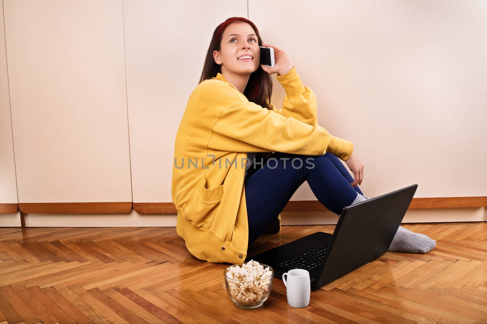 Communication, friendship concept. Girl talk concept. Beautiful young woman sitting on the floor in her room in front of her laptop with coffee and popcorn, and talking on the mobile phone.