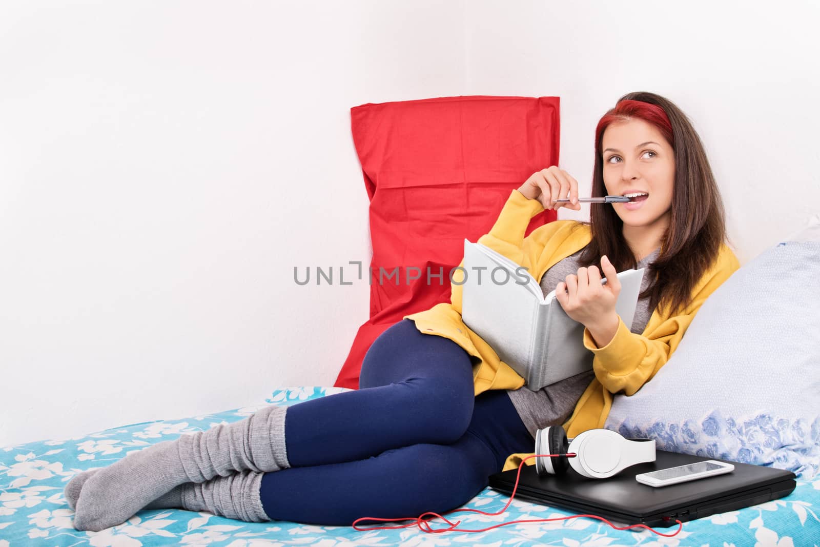 Beautiful young girl in her room holding a notebook, biting a pen, thinking, wondering, while sitting in bed next to laptop and headphones. Girl writing in her diary.