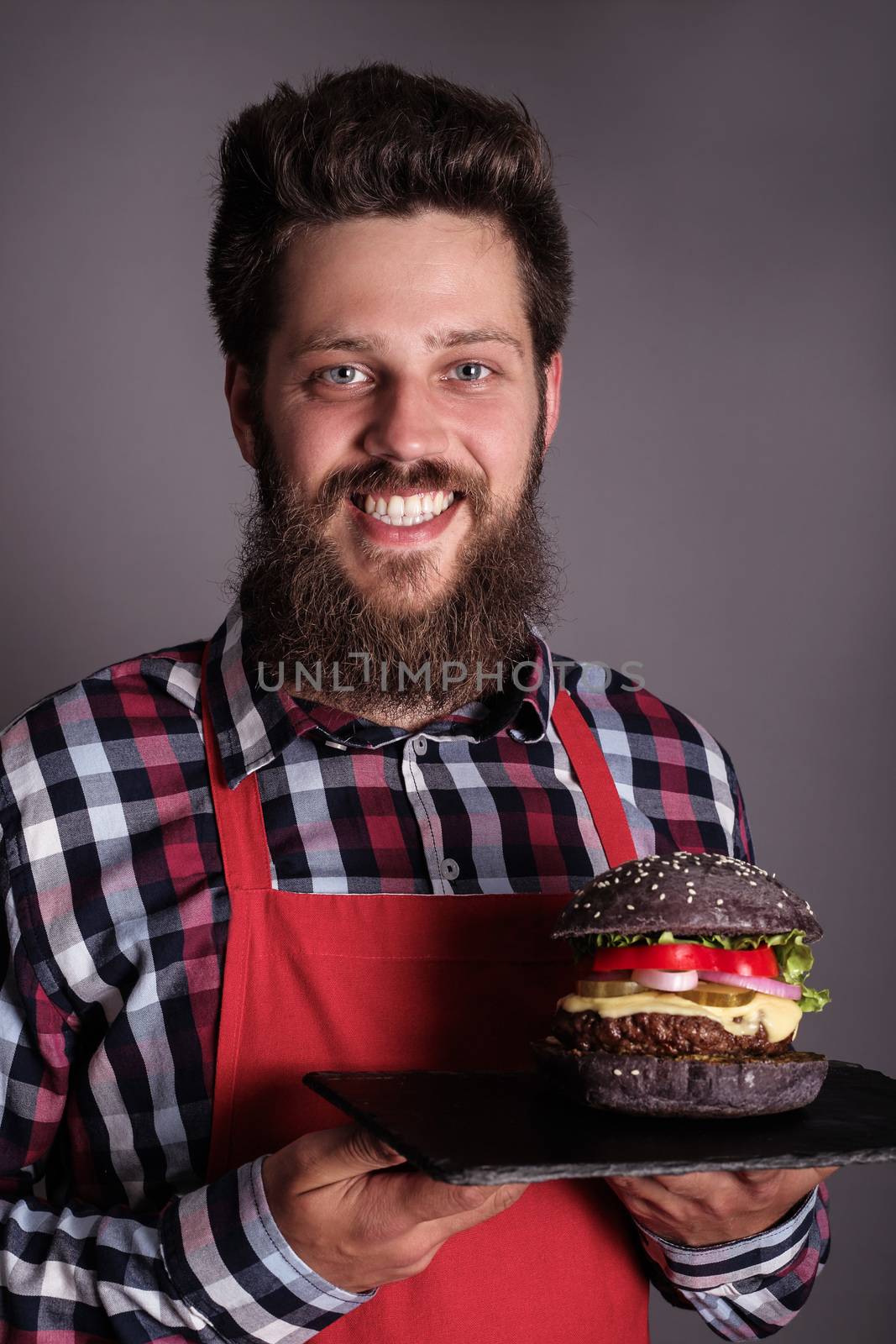 Happy smiling male cook in red apron proudly presents fresh self made black burger , gray background