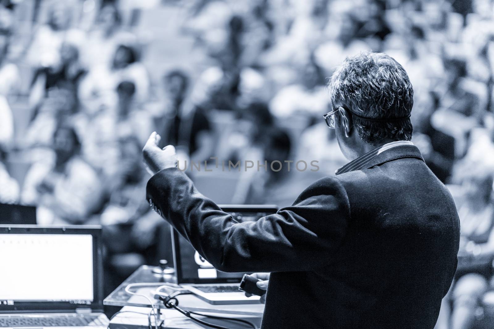 Speaker giving a talk on corporate business conference. Unrecognizable people in audience at conference hall. Business and Entrepreneurship event. Black and white, blue toned image.