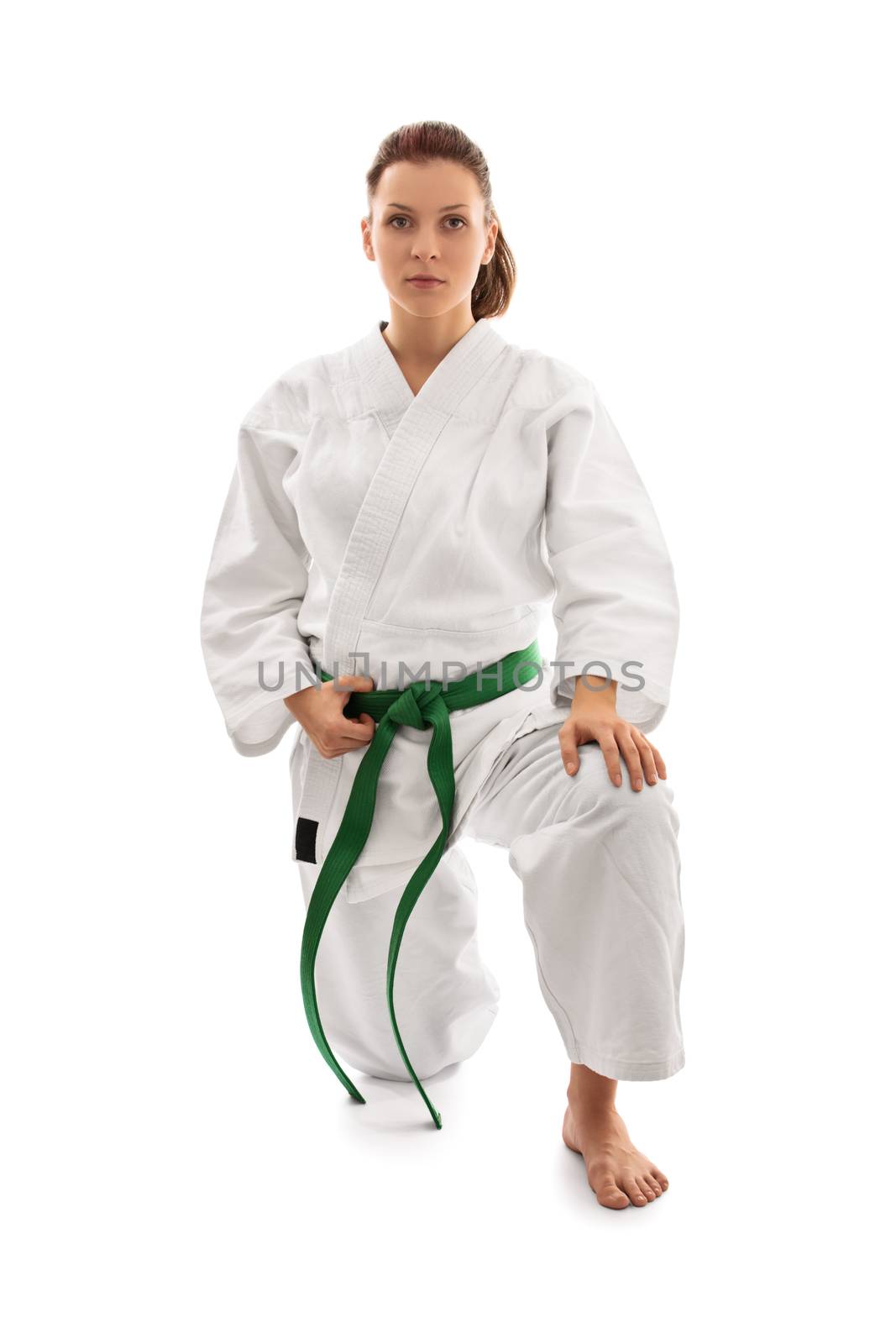Young female martial arts fighter in white kimono and green belt kneeling on one knee and looking serious, isolated on white background. Body and spirit bent to my will.