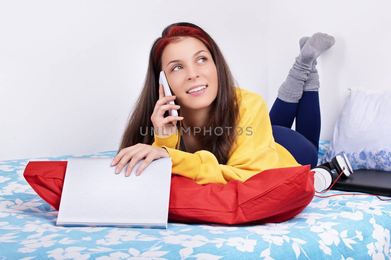 Communication, friendship concept. Beautiful young woman lying on the bed in her room with feet in the air talking on the mobile phone. Young girl on the phone talking about a good book.
