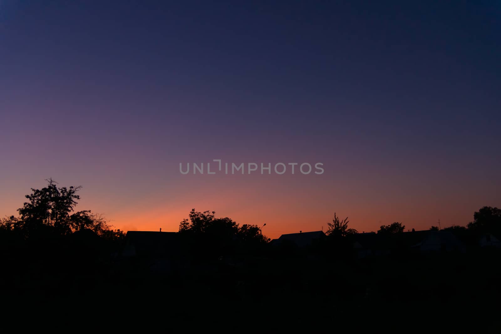 Evening sunset in the village a far away from the city. The sky is saturated with orange and blue colours. On foreground silhouttes of trees. Underexposed low light photo.