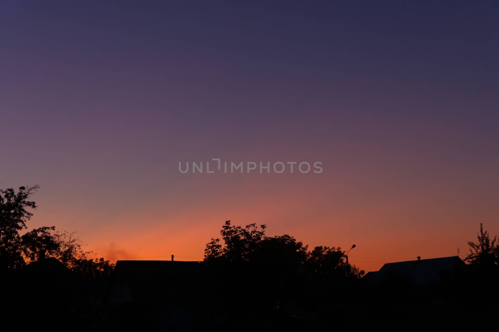 Evening sunset in the village a far away from the city. The sky is saturated with orange and blue colours. On foreground silhouttes of trees. Underexposed low light photo.