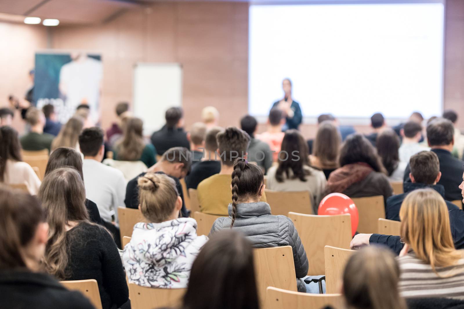 Business and entrepreneurship symposium. Female speaker giving a talk at business meeting. Audience in conference hall. Rear view of unrecognized participant in audience. Copy space on whitescreen.