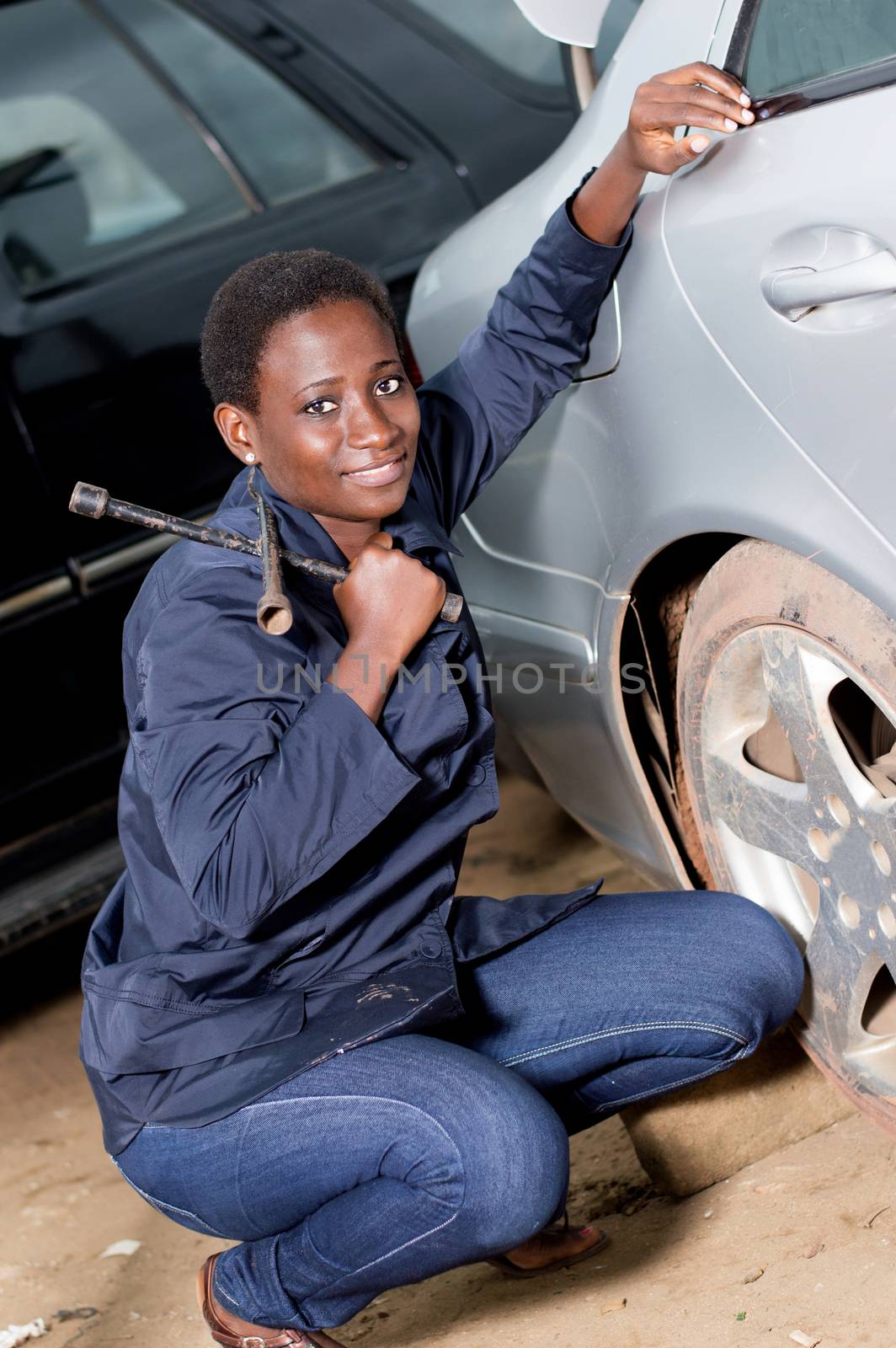 Young smiling mechanic squatting near the tire of a car she is preparing to remove.
