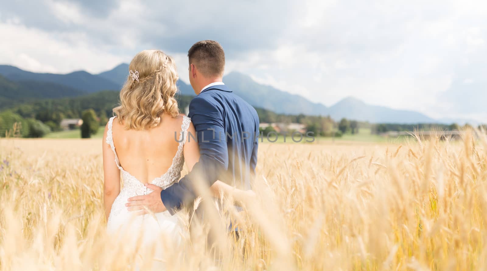 Rear view of groom huging bride tenderly in wheat field somewhere in Slovenian countryside. Caucasian happy romantic young couple celebrating their marriage.