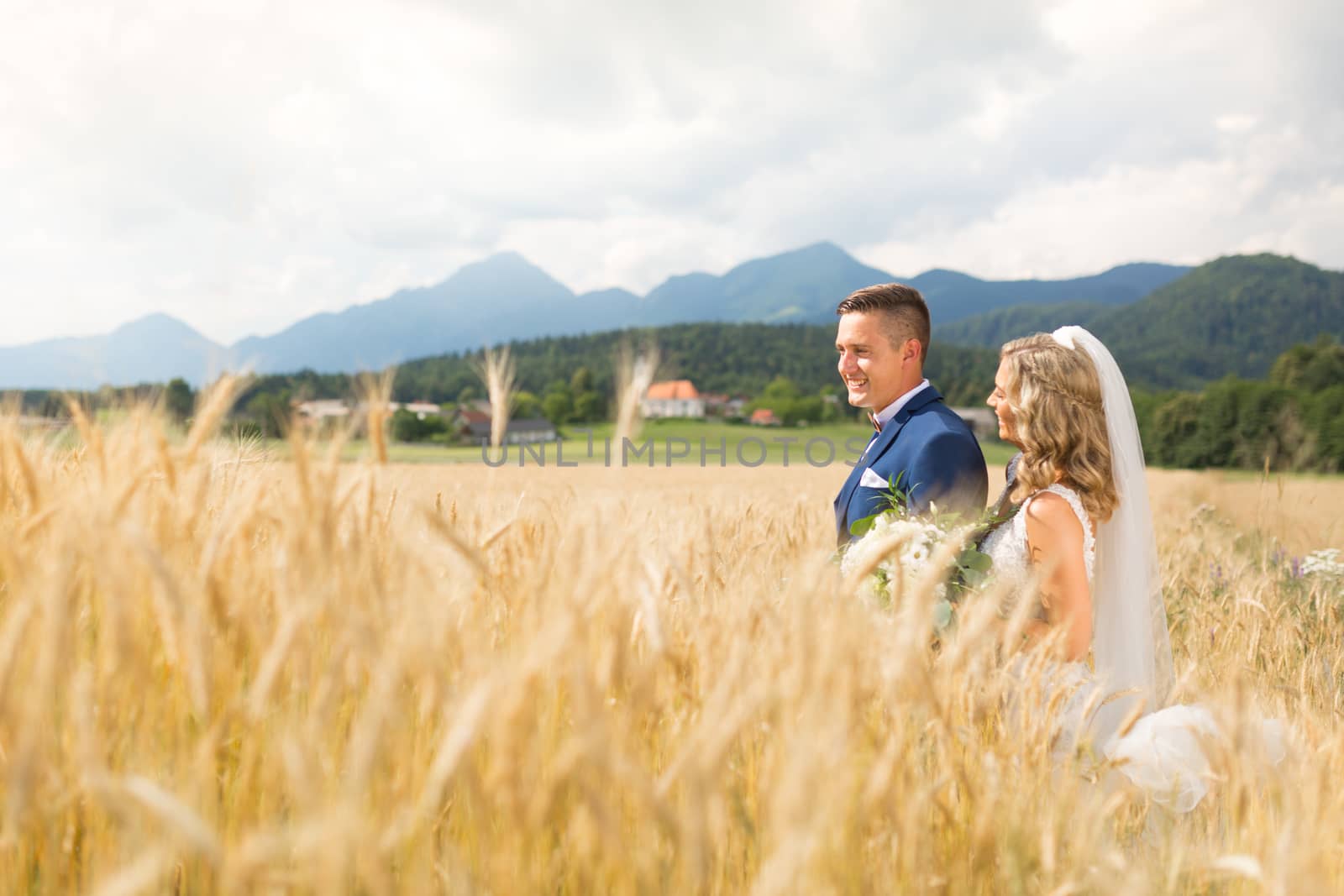 Bride hugs groom tenderly in wheat field somewhere in Slovenian countryside. Caucasian happy romantic young couple celebrating their marriage.