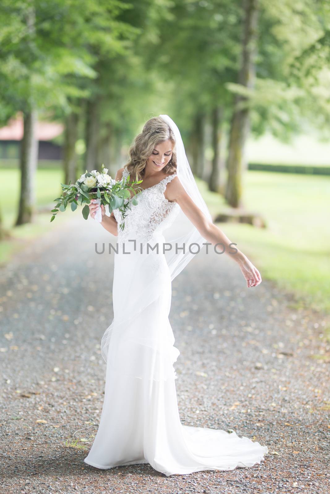 Full length portrait of beautiful sensual young blond bride in long white wedding dress and veil, holding bouquet outdoors in natural background by kasto