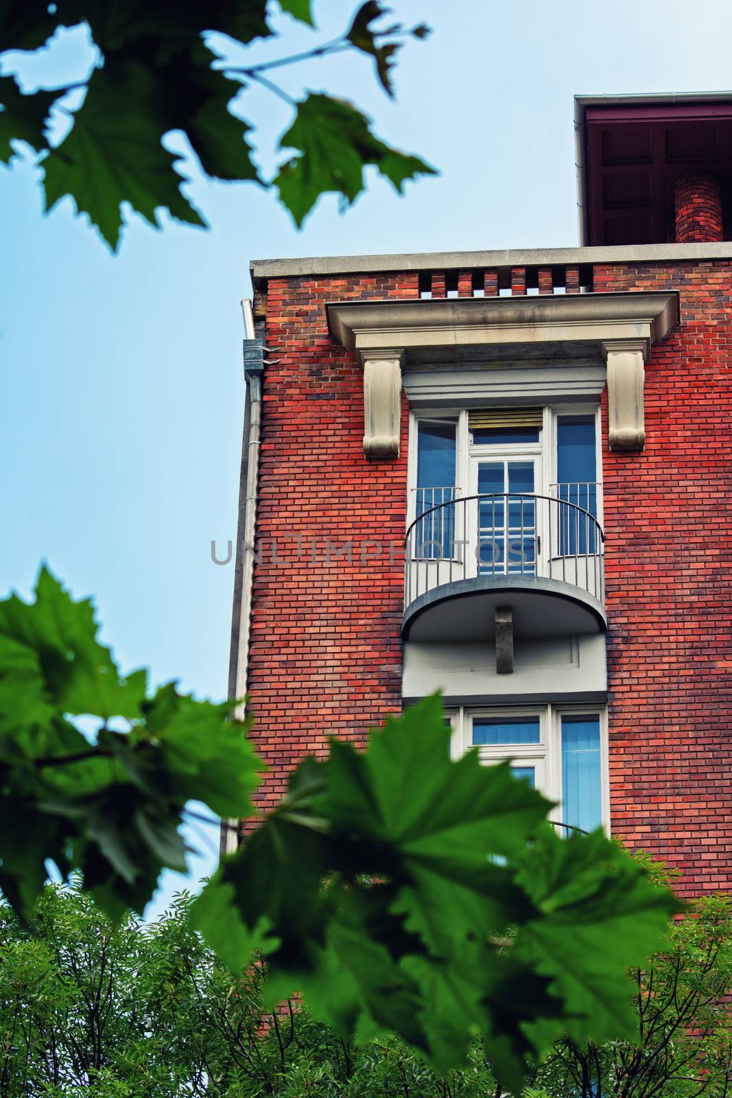 Low angle view of a balcony on a beautiful residential building with red brick framed by tree branches and green leaves.
