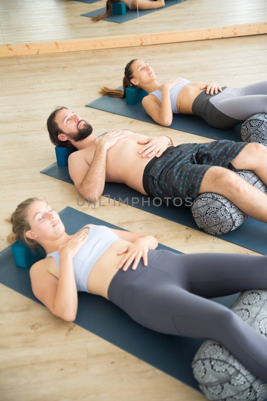 Restorative yoga with a bolster. Group of young sporty attractive people in yoga studio, lying on bolster cushion, stretching and relaxing during restorative yoga. Healthy active lifestyle.