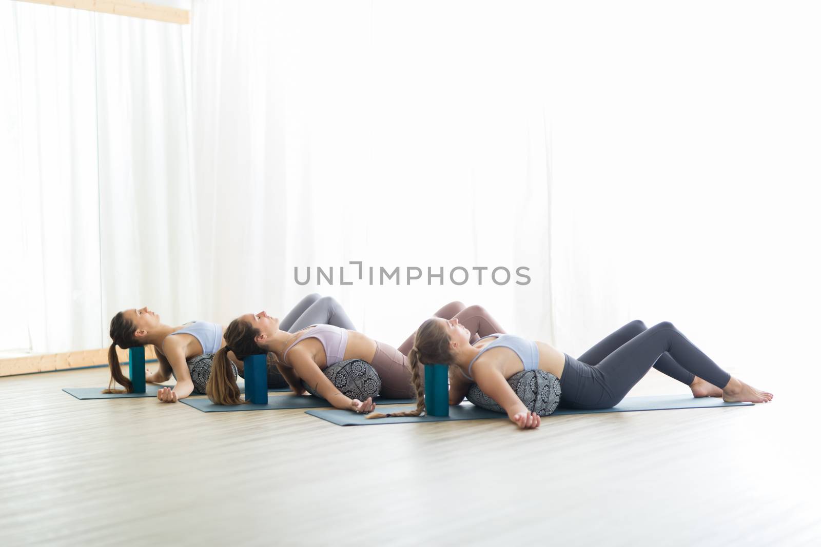 Restorative yoga with a bolster. Group of three young sporty attractive women in yoga studio, lying on bolster cushion, stretching and relaxing during restorative yoga. Healthy active lifestyle by kasto
