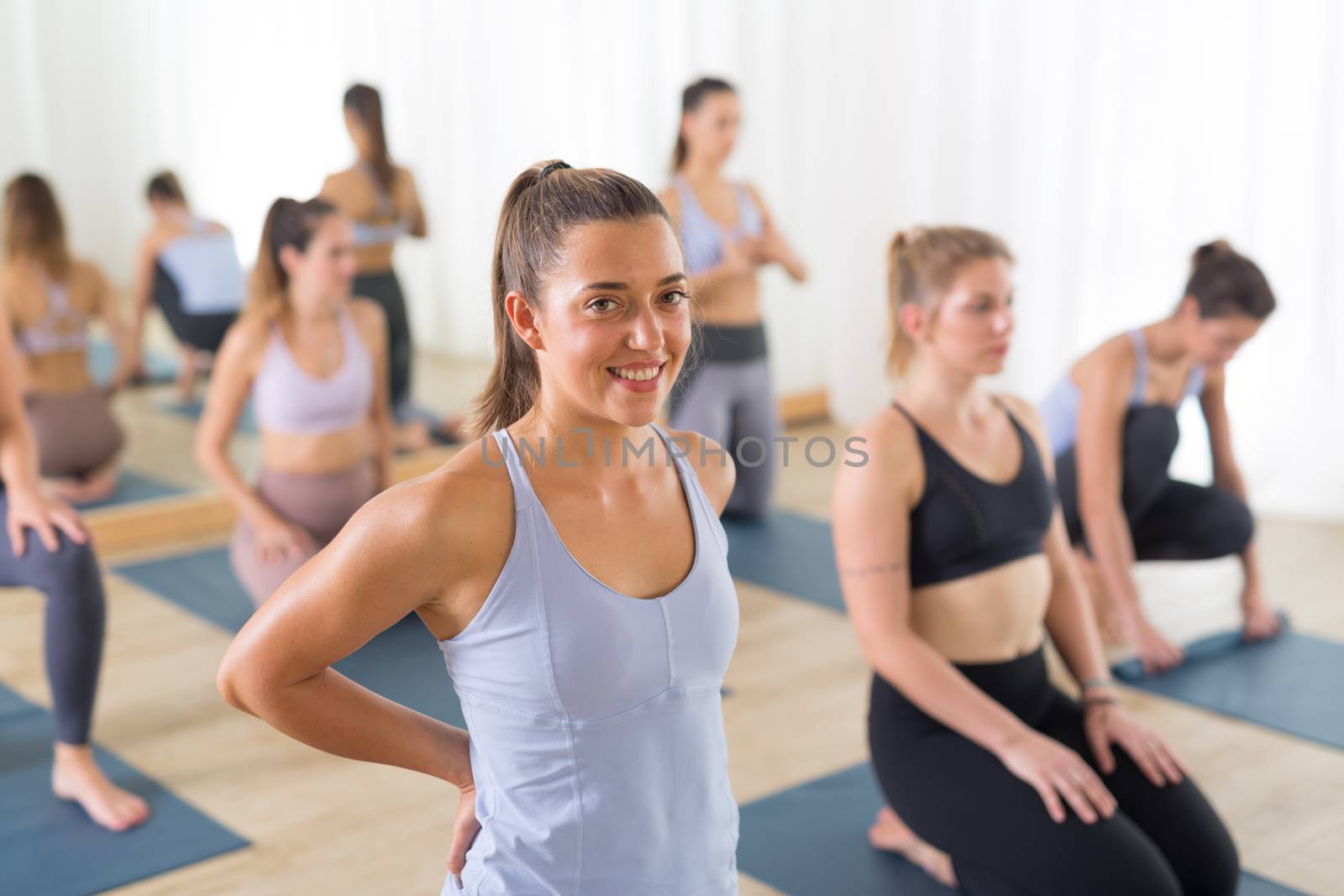 Portrait of a cheerful beautiful young yoga instructor relaxing after giving yoga class to large group of sporty attractive people. Healthy active lifestyle, working out indoors in gym. by kasto
