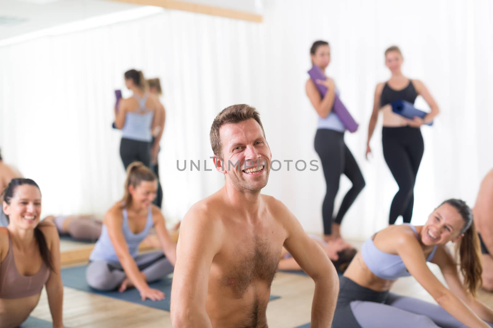 Portrait of a cheerful beautiful young yoga instructor relaxing after giving yoga class to a large group of attractive sporty people. Healthy active lifestyle, working out indoors in gym.