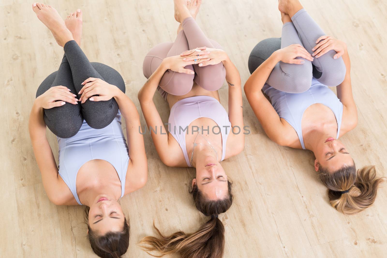 Group of three young sporty attractive women in yoga studio, lying on the floor, stretching and relaxing after the workout. Healthy active lifestyle, working out indoors in gym by kasto