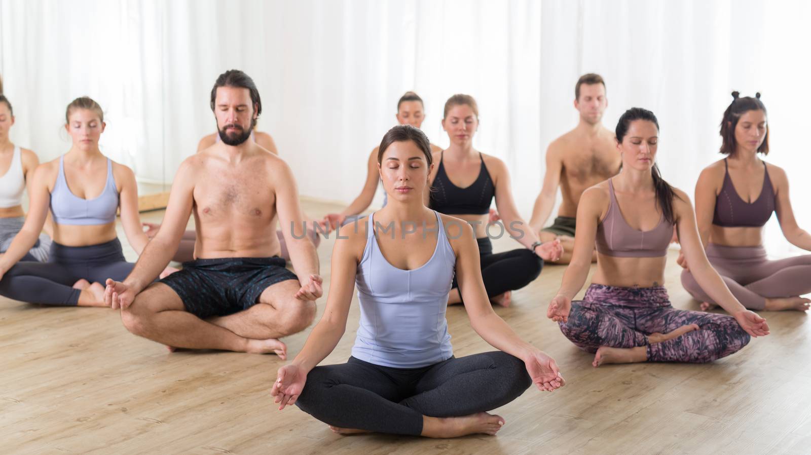 Group of young sporty attractive people in yoga studio, practicing yoga lesson with instructor, sitting on floor in Siddhasana, easy seated yoga pose. Healthy active lifestyle, working out in gym by kasto