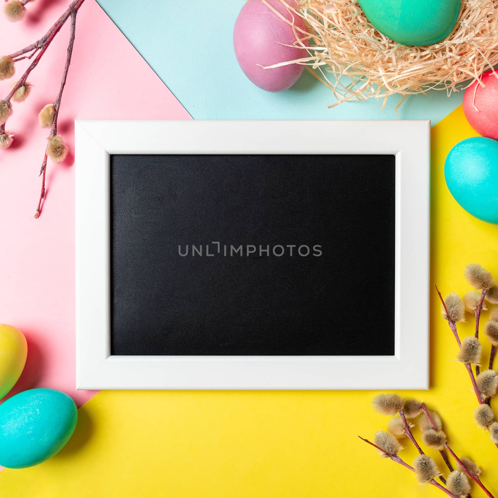 Orthodox Easter concept. Colorful eggs and pussy willow branches on bright colorful background with empty chalkboard. Copy space for greetings, text or design. Top down view or flat lay. Square crop