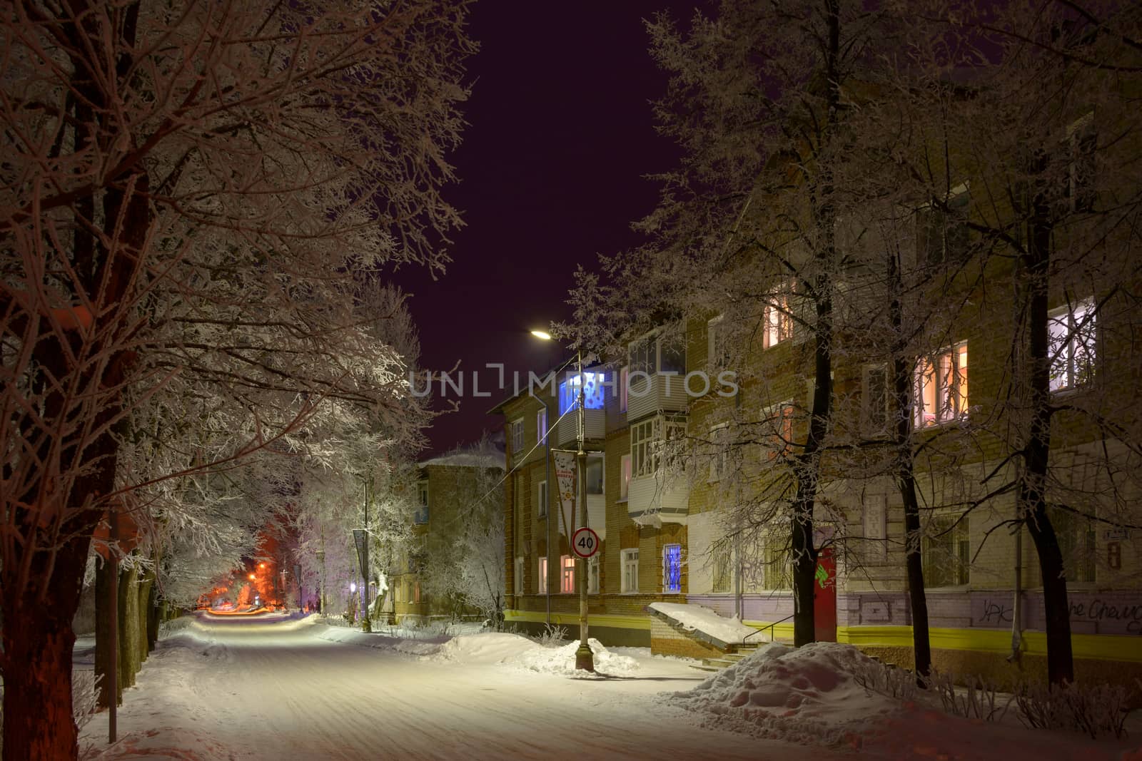 Night snowy street with trees in the light of lanterns and windows of houses painted with different colors.