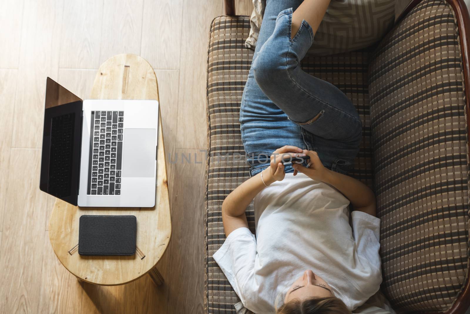 Woman use a smartphone while lying on a sofa with a laptop and ereader on a table. Studying online, freelance. Self employed or freelance girl use a phone and resting from work whith a notebook