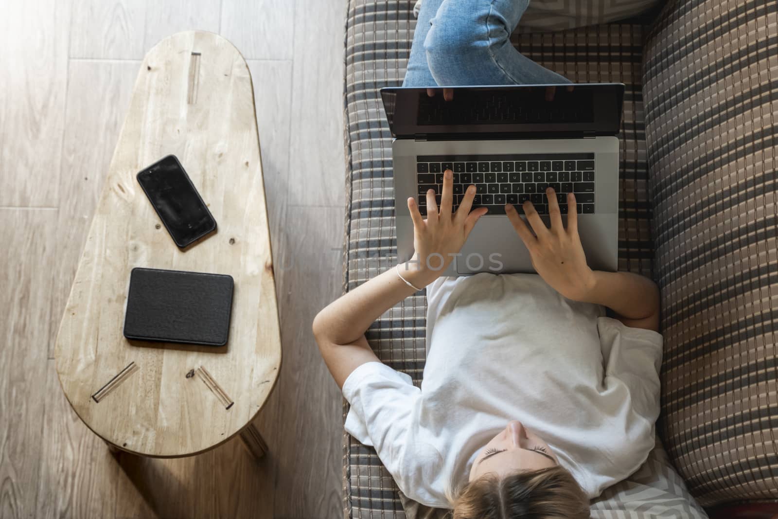 Woman with a laptop laying on a sofa. Study and work online, freelance. Self employed woman, girl working with her notebook laying on a couch with a phone, smartphone and ereader on table