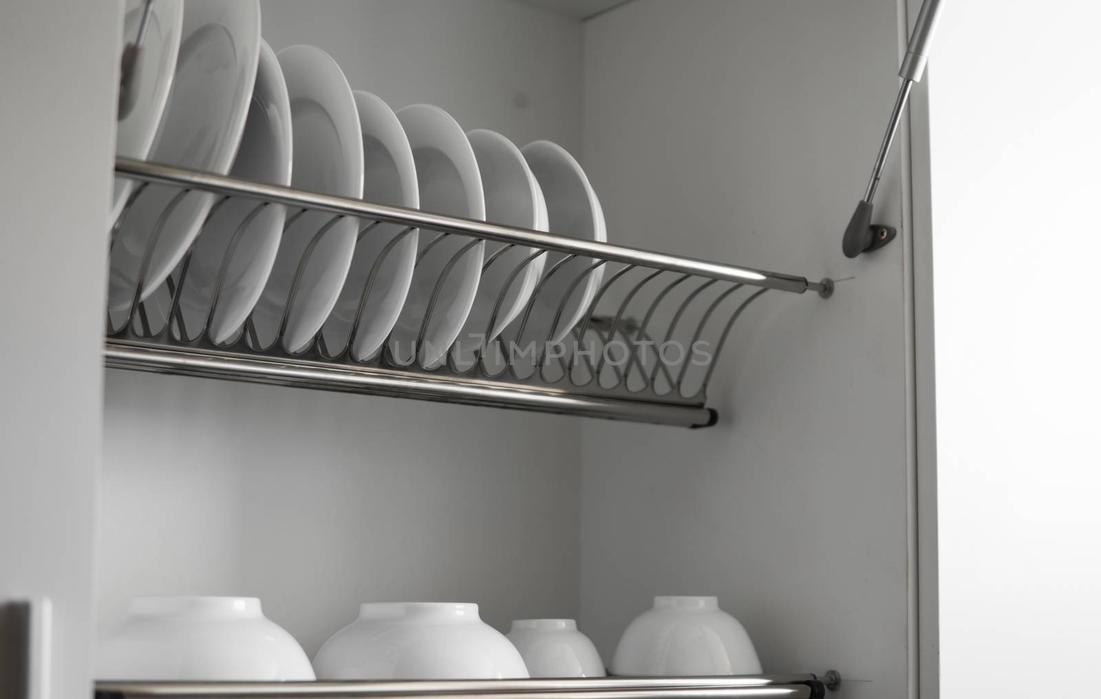 Dish drying metal rack with big nice white clean plates. Traditional comfortable kitchen. Open white dish draining closet with wet dishes of glass and ceramic, plates, bowls drying inside on rack