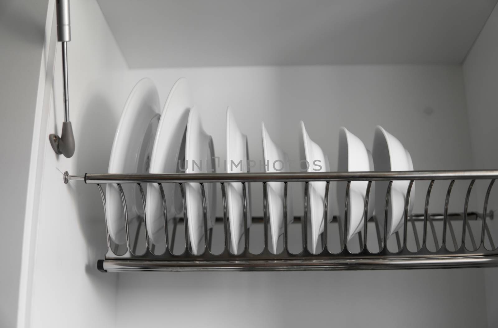 Dish drying metal rack with big nice white clean plates. Traditional comfortable kitchen. Open white dish draining closet with wet dishes of glass and ceramic, plates, bowls drying inside on rack