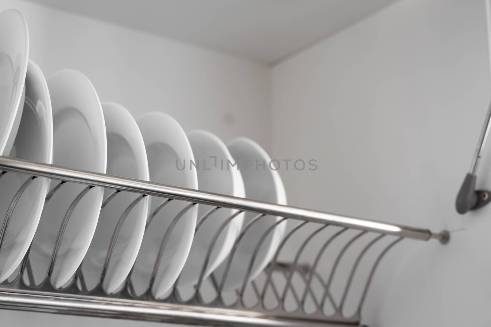 Dish drying metal rack with big nice white clean plates. Traditional comfortable kitchen. Open white dish draining closet with wet dishes of glass and ceramic, plates, bowls drying inside on rack. by vovsht