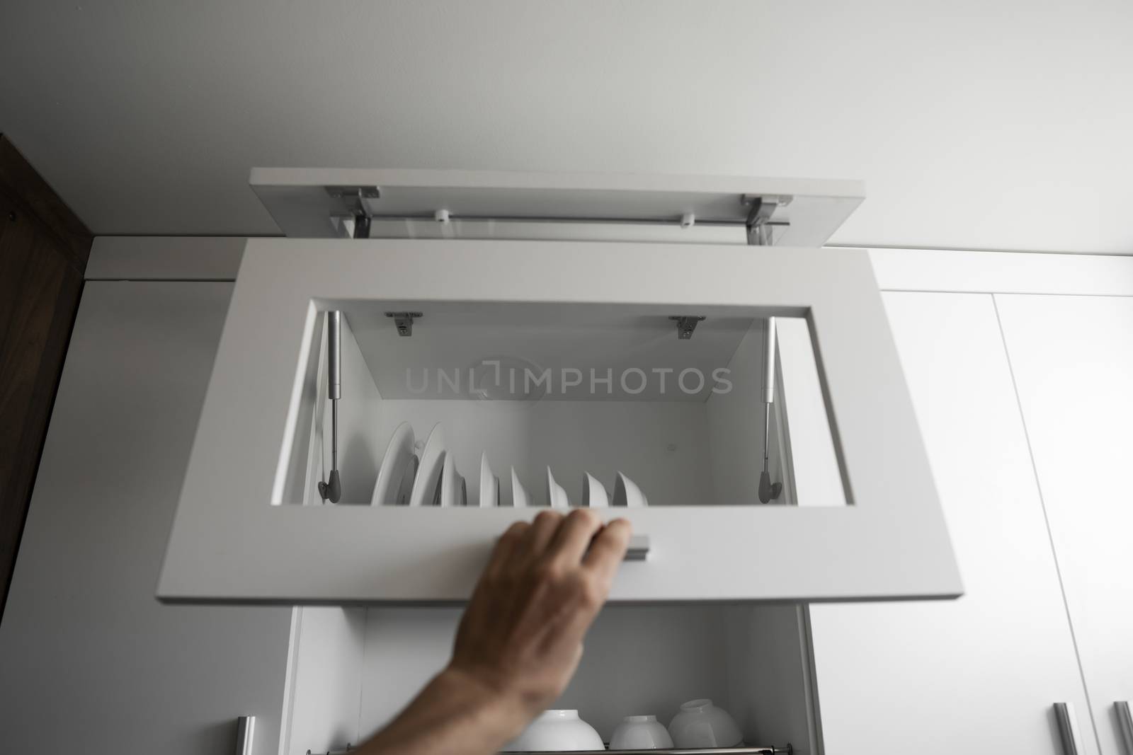 Hand holds the door of the dryer with utensils. Dish drying metal rack with white clean plates. Traditional comfortable kitchen. Open white dish draining closet with wet dishes of plates, bowls. by vovsht