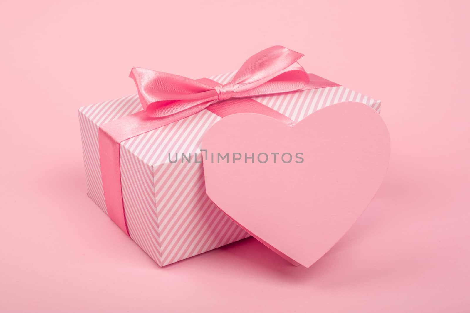 Valentine Day gift in a box wrapped in striped paper and tied with silk ribbon bow and heart shapes greeting card on pink background with copy space for text