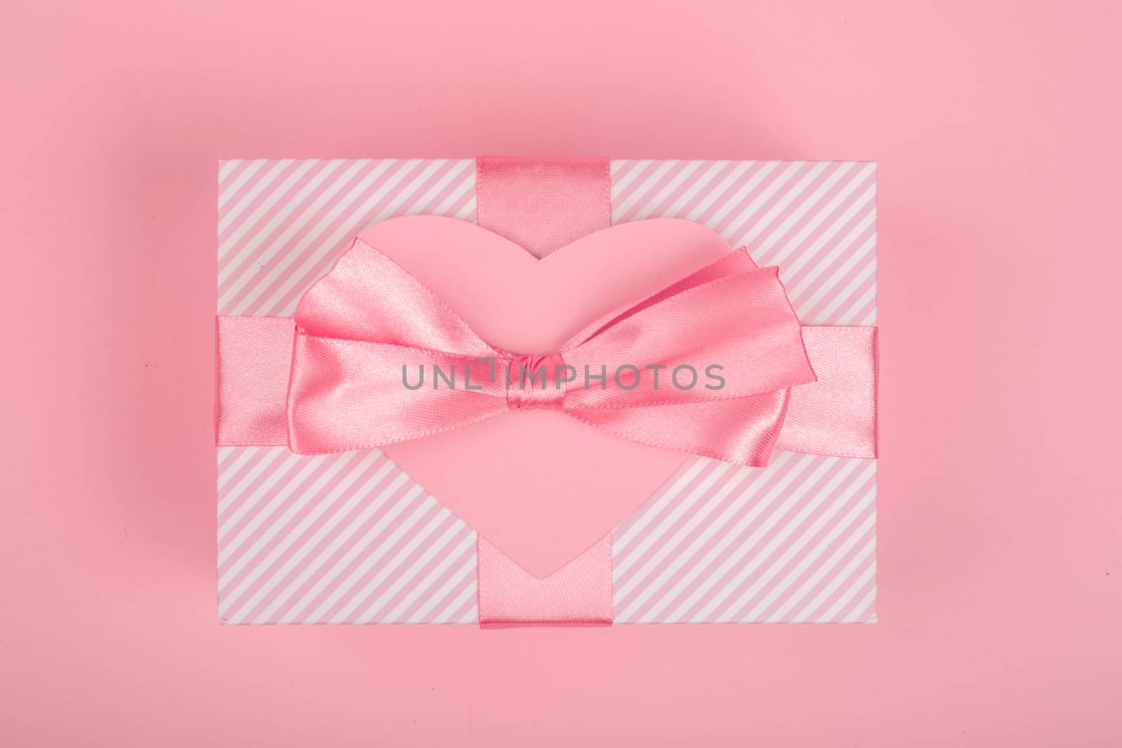 Valentine Day gift in a box wrapped in striped paper and tied with silk ribbon bow and heart shapes greeting card on pink background with copy space for text