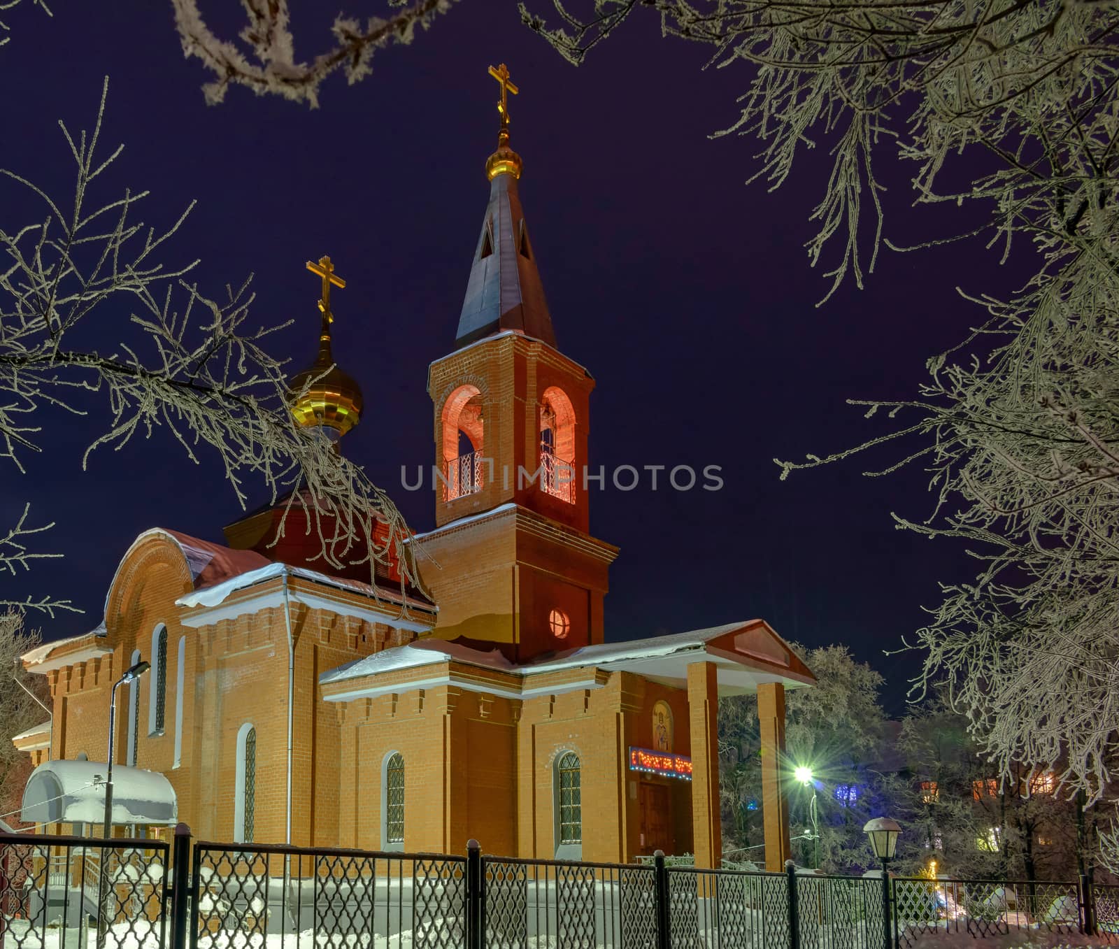 The building of the Orthodox Church at night surrounded by trees in the snow illuminated by lanterns.