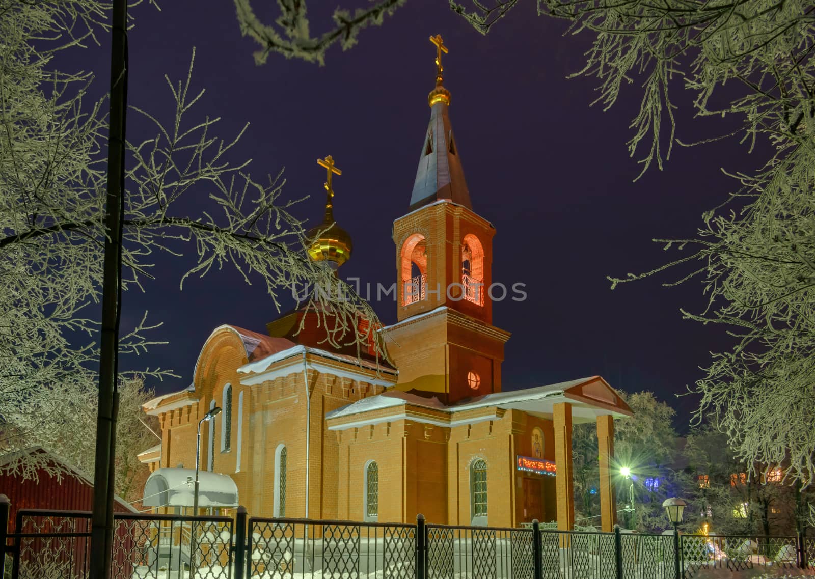 The building of the Orthodox Church at night surrounded by trees in the snow illuminated by lanterns.